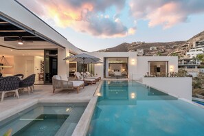 A modern outdoor pool area with lounge chairs and a dining setup, surrounded by a white building with a view of the hills.