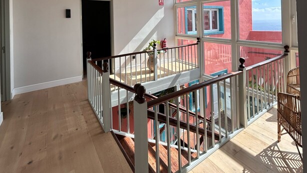 A spacious room with a high ceiling, a staircase leading to an upper level, and large windows offering a view of a red building.