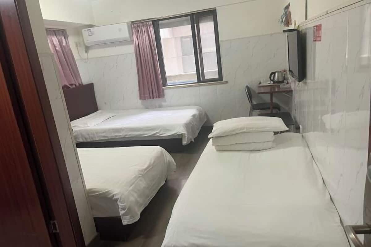 A room with three single beds, a desk with a chair, an air conditioning unit, and a window with curtains.
