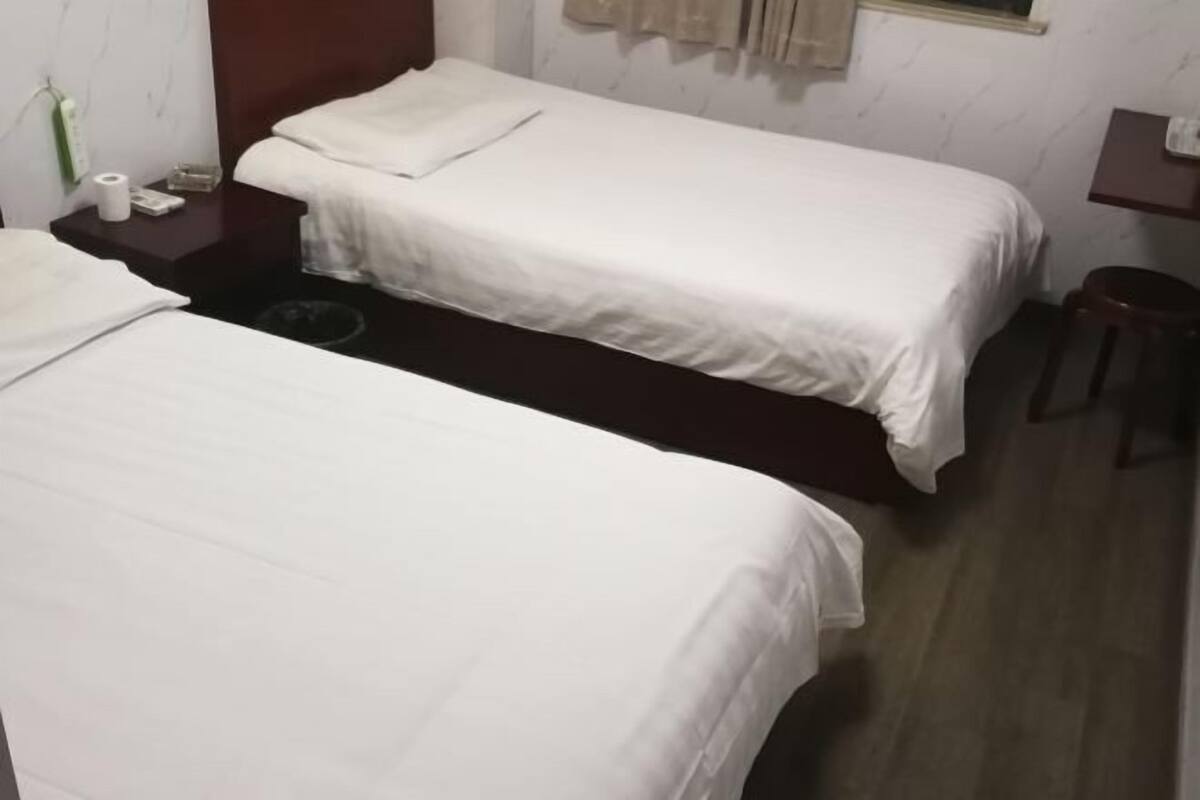 A hotel room with two single beds, a small table, a chair, and a nightstand with a lamp and some items on it.