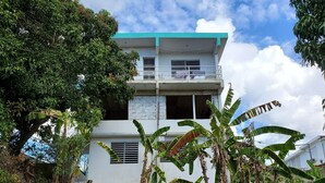 A two-story building with a balcony, surrounded by banana trees and a garden area. - Beautiful home with 2 bedrooms 2 minutes from the beach. (Rincon)