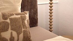 Una cama bien hecha con edredón color crema y almohadas estampadas, una lámpara de mesa redonda y un radiador en una habitación con paredes texturizadas.