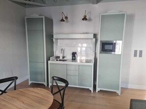 Cocina compacta con lavamanos, microondas y cafetera. Hay dos sillas y una mesa redonda.