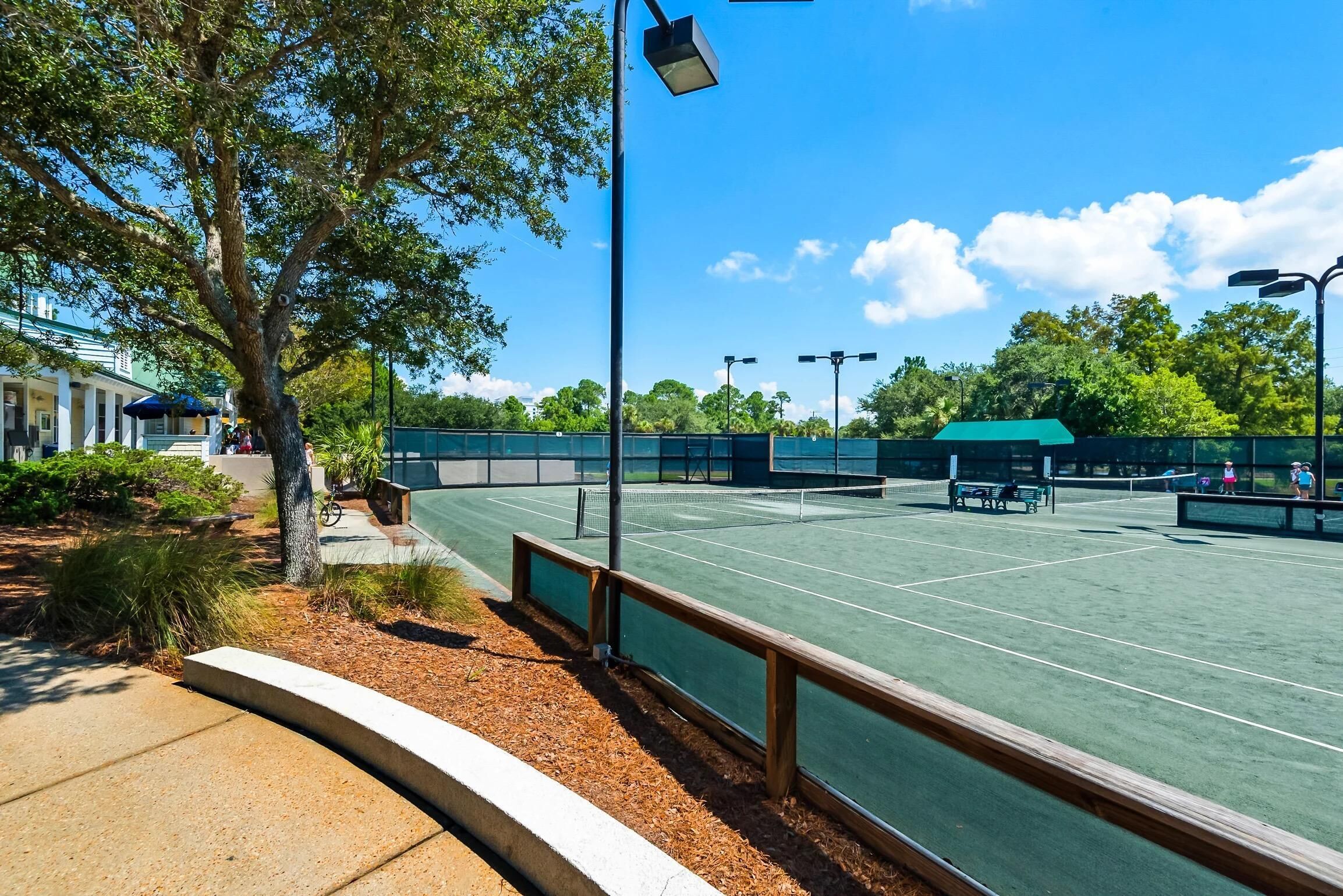 An outdoor tennis court with a wooden fence, trees, and a clear sky.