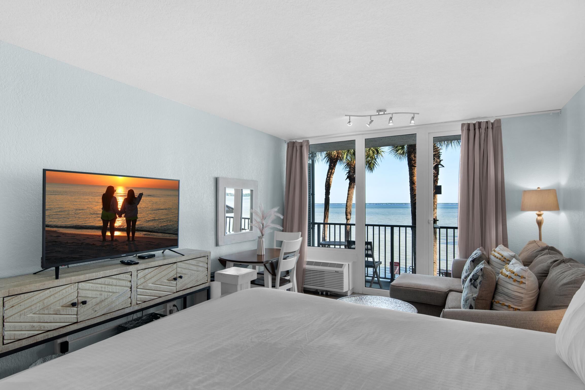 Hotelkamer met een groot bed, een tv waarop een zonsondergang te zien is, een balkon met uitzicht op de zee en een zithoek met een tafel en stoelen.