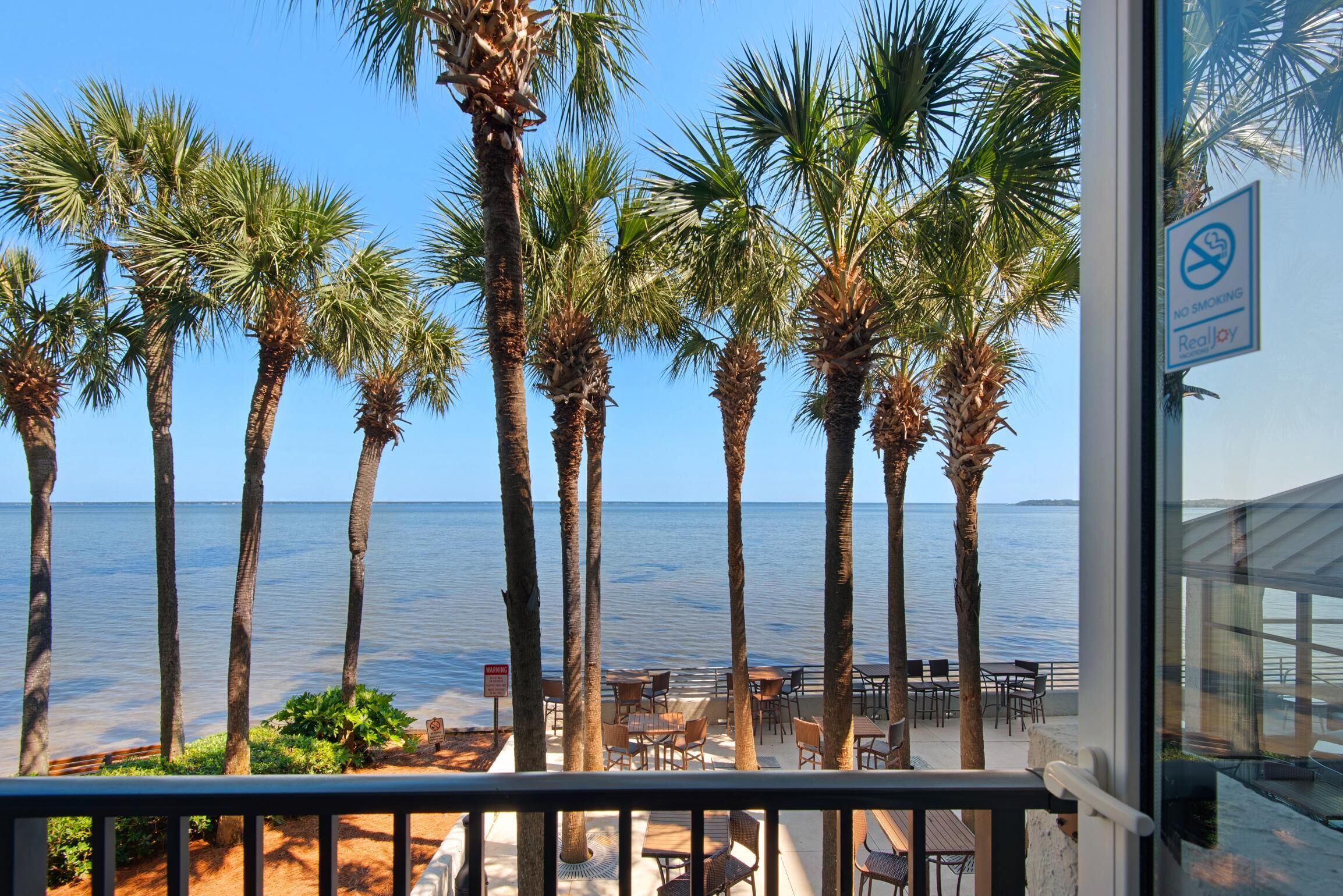 Vista de un restaurante frente a la playa con mesas al aire libre, palmeras y una vista despejada del océano.