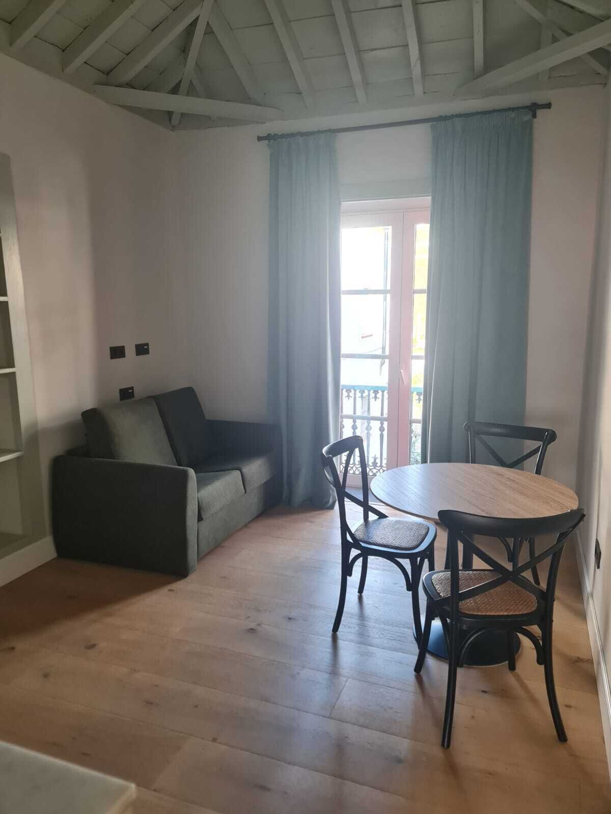 A room with a wooden floor, a grey sofa, a round table with four chairs, and a window with blue curtains.