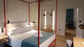 Ein Bett mit Baldachin, mit rotem Gestell, weißer Bettwäsche und einer blauen Decke. Ein Holztisch mit Stuhl, einer Lampe und einem kleinen Tisch ebenfalls mit Lampe. Der Raum hat Holzdielen und eine hohe Decke mit freiliegenden Balken.