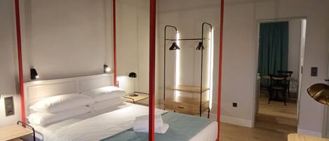 Un lit à baldaquin avec un cadre rouge, du linge de lit blanc et un plaid bleu. On y trouve un bureau en bois avec une chaise, une lampe et une petite table avec une lampe. La pièce est dotée d’un plancher en bois et d’un plafond haut avec des poutres apparentes.
