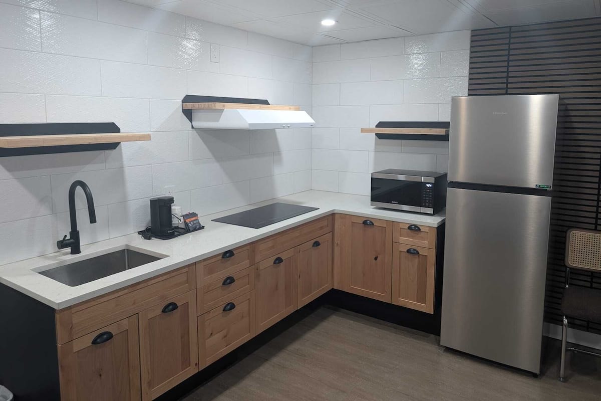 Una cocina moderna con paredes de azulejos blancos, armarios de madera, un refrigerador de acero inoxidable, un microondas y una cafetera.