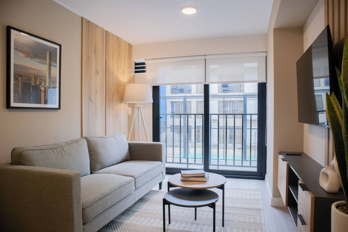Ein modernes Wohnzimmer mit einer Couch, einem kleinen Tisch mit Büchern, einer Stehlampe und einem großen Fenster mit Blick auf Gebäude.