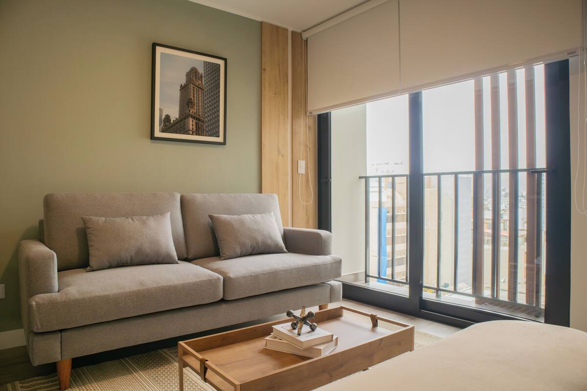 Ein modernes Wohnzimmer mit einem grauen Sofa, einem Couchtisch mit Büchern, einem gerahmten Bild an der Wand und einem großen Fenster mit Blick auf Gebäude.