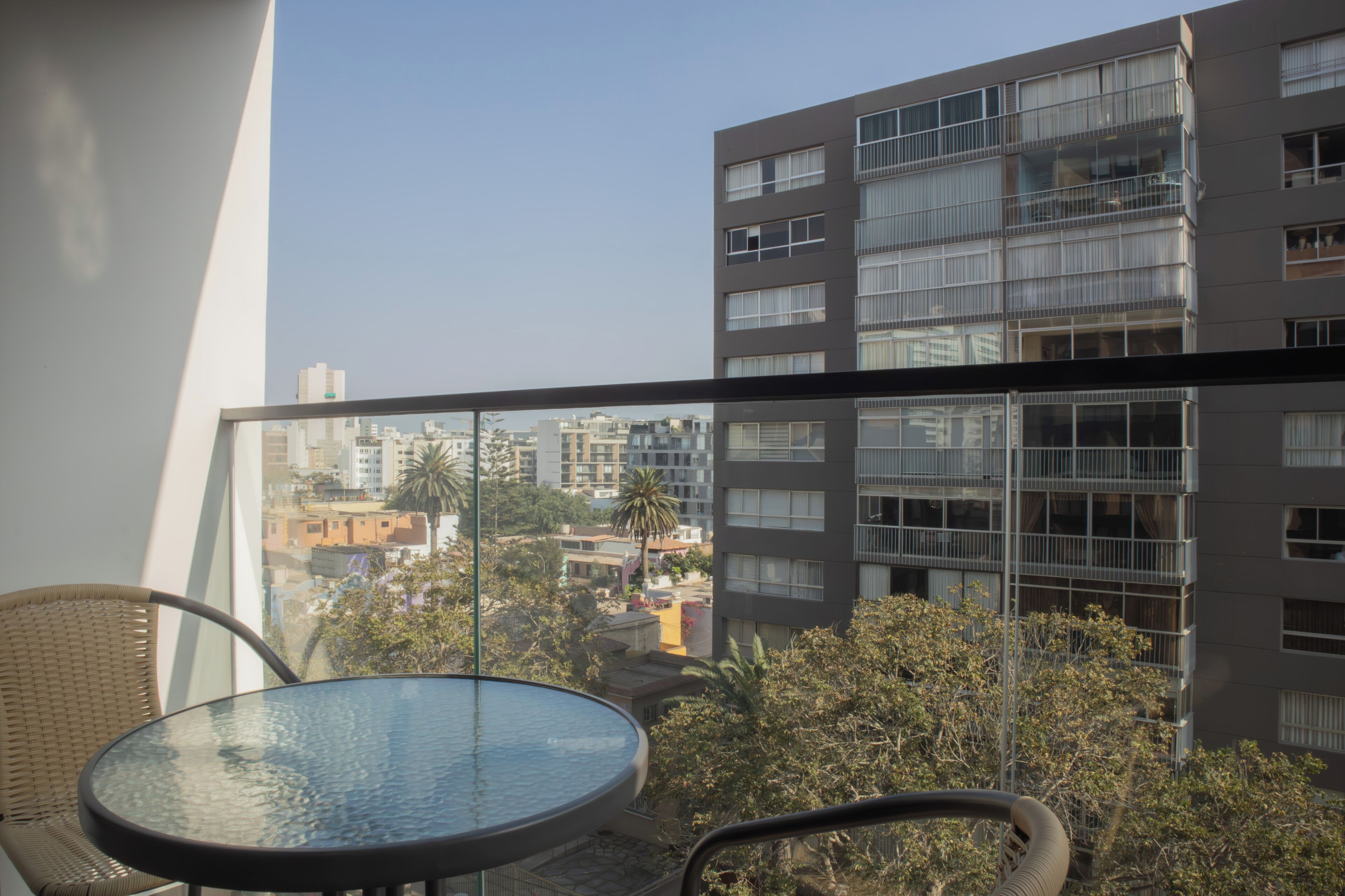 A balcony with a glass table and wicker chairs, overlooking a cityscape with modern buildings.