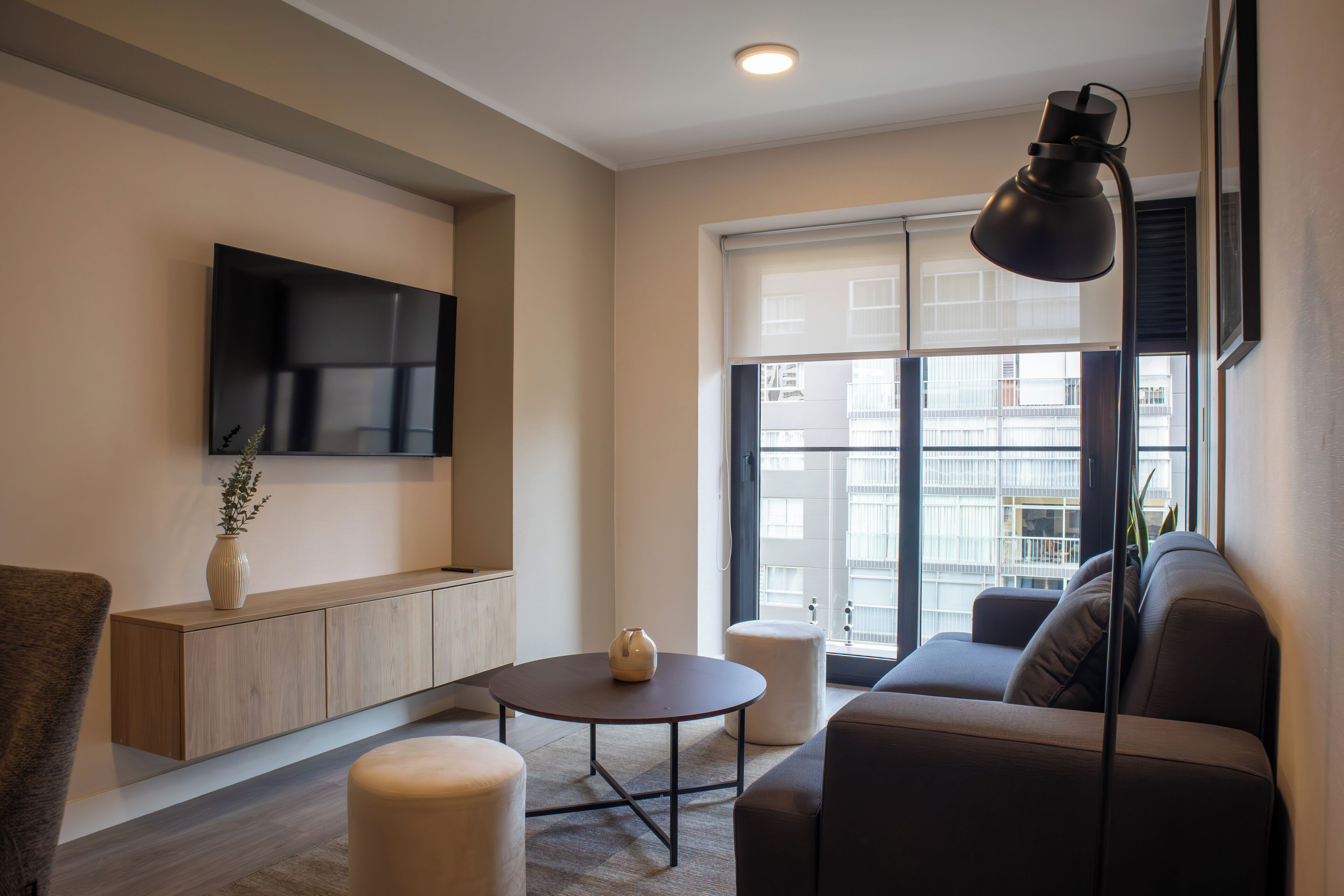 A modern living room with a sofa, a coffee table, a TV mounted on the wall, and a window with a view of buildings.