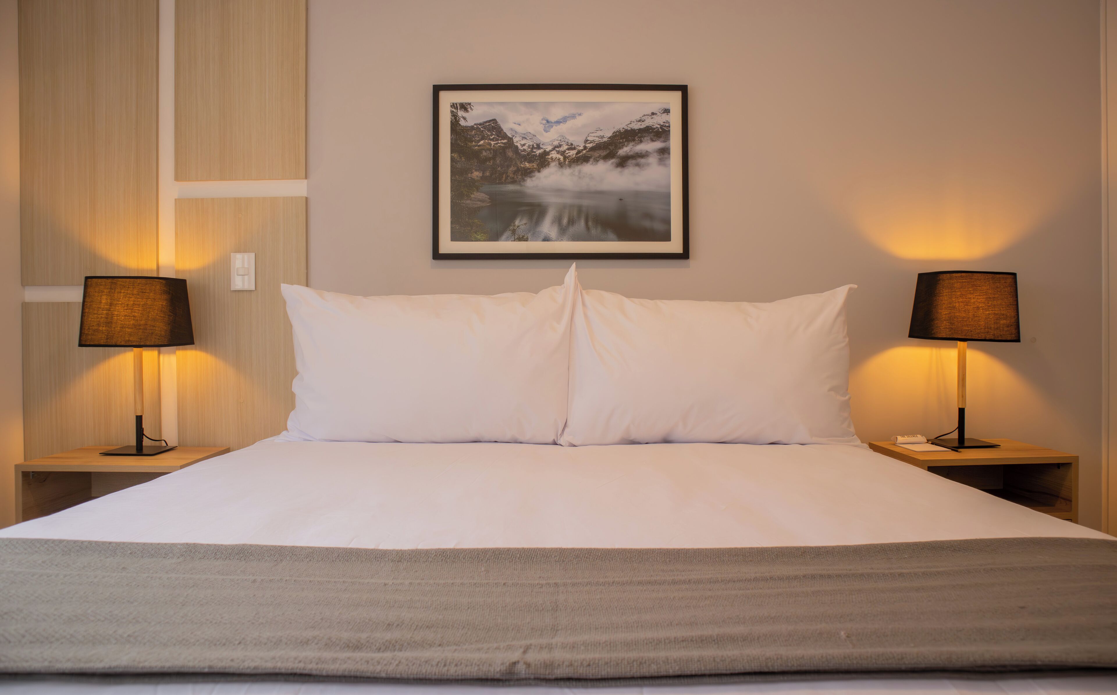 Una cama bien hecha con sábanas blancas, dos lámparas de noche y un cuadro enmarcado con un paisaje montañoso en la pared.