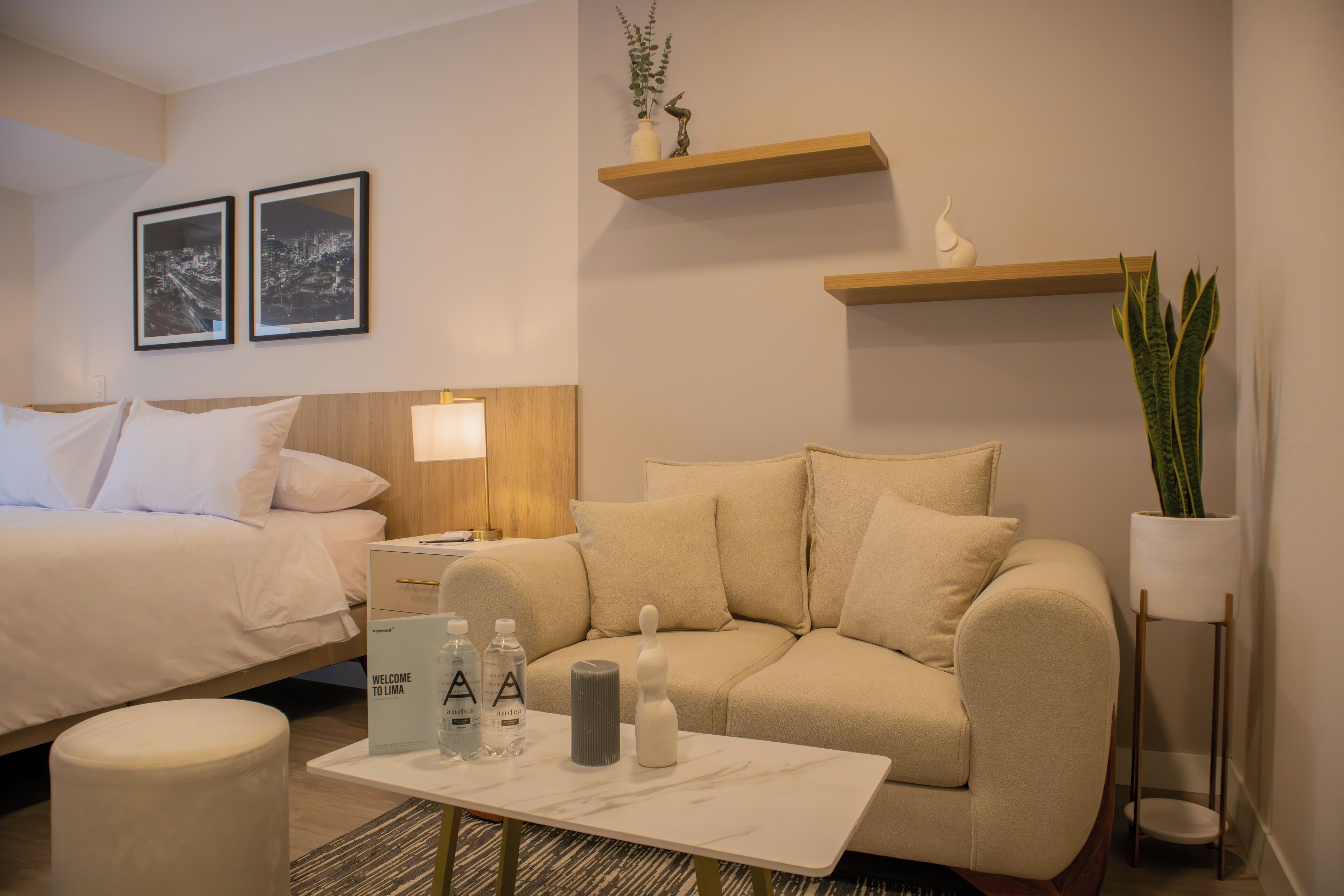 A hotel room with a bed, a sofa, a nightstand, a table, and two wall-mounted shelves with decorative items.