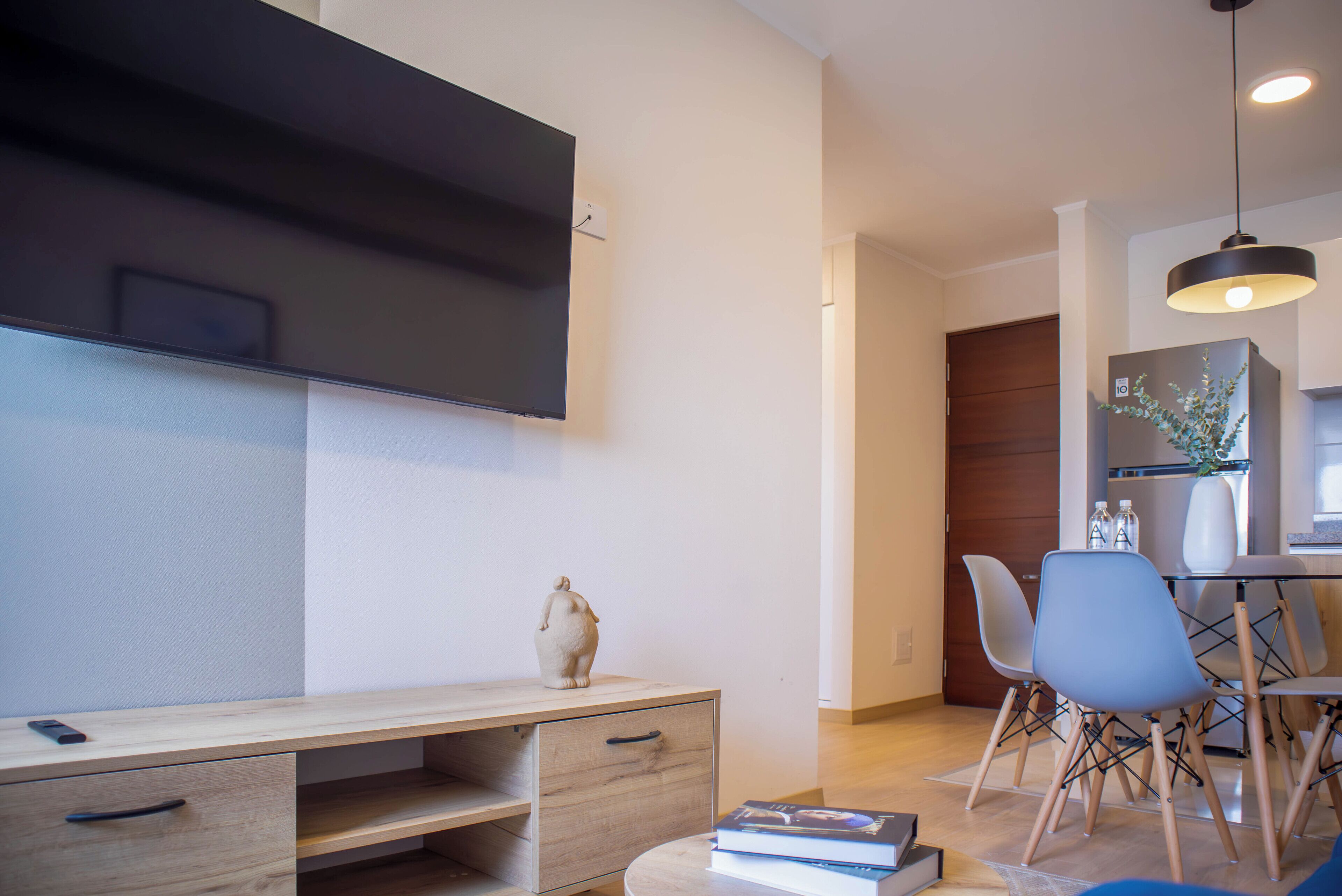 Un salon moderne avec un téléviseur à écran plat fixé au mur, un bureau en bois avec des tiroirs, un vase et un coin repas comprenant des chaises et une table.