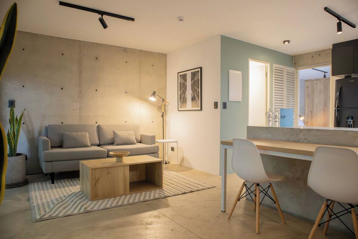 Ein modernes Wohnzimmer mit Sofa, Couchtisch und Sesseln, ein Küchenbereich mit Arbeitsplatte und Stühlen sowie eine Wand mit Betonoptik.