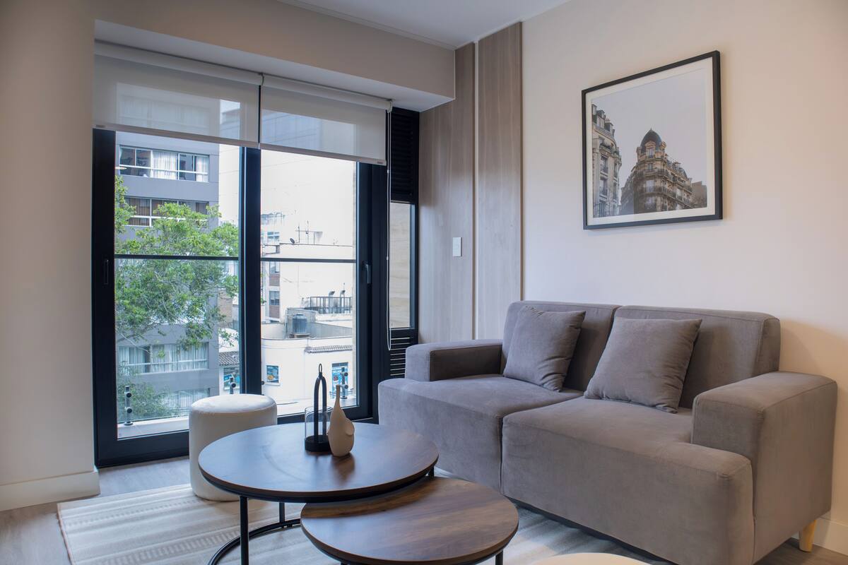 Ein modernes Wohnzimmer mit einem grauen Sofa, einem runden Holztisch und einem großen Fenster mit Blick auf Gebäude und Bäume.
