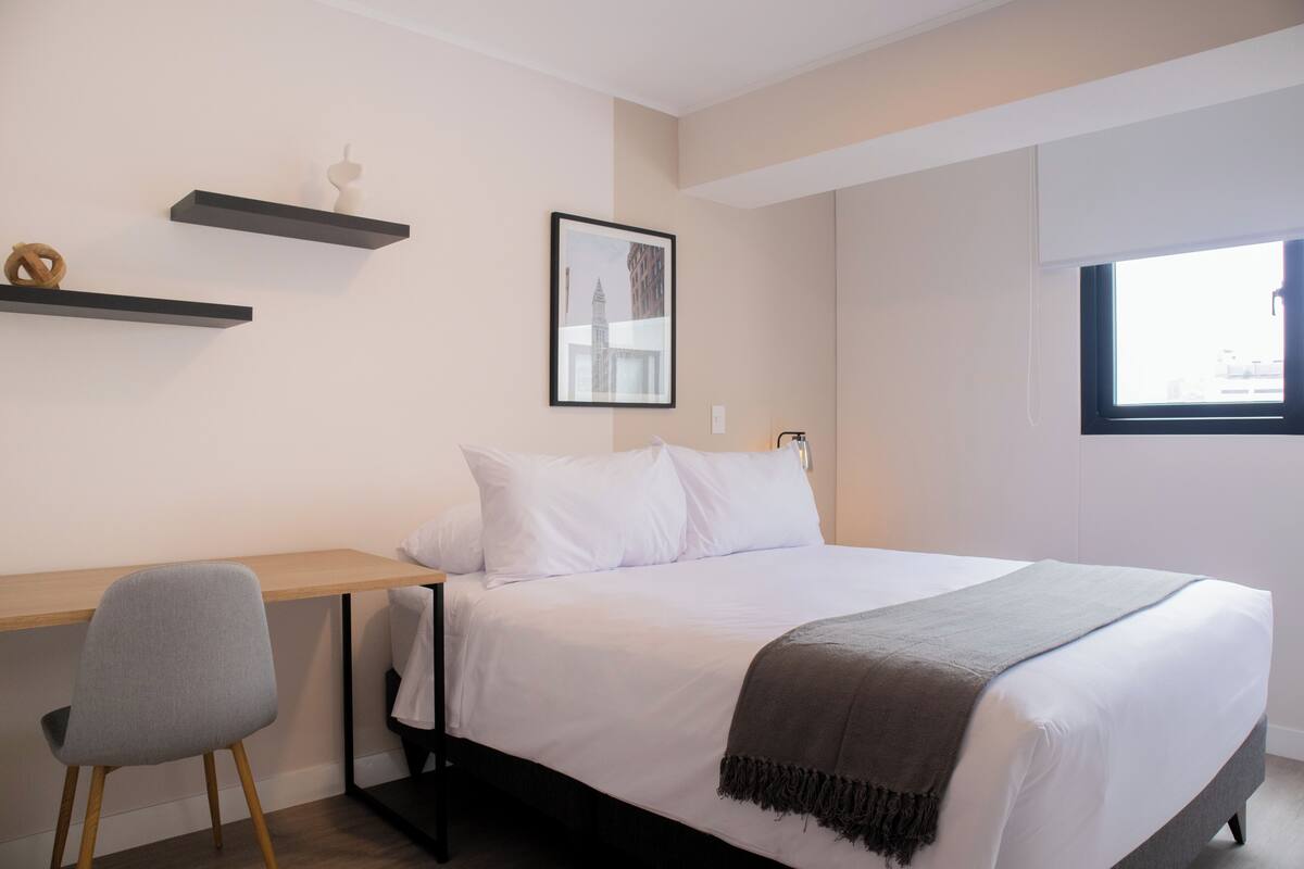 Ein Schlafzimmer mit Bett, Schreibtisch, Stuhl, Fenster und einem gerahmten Bild an der Wand.