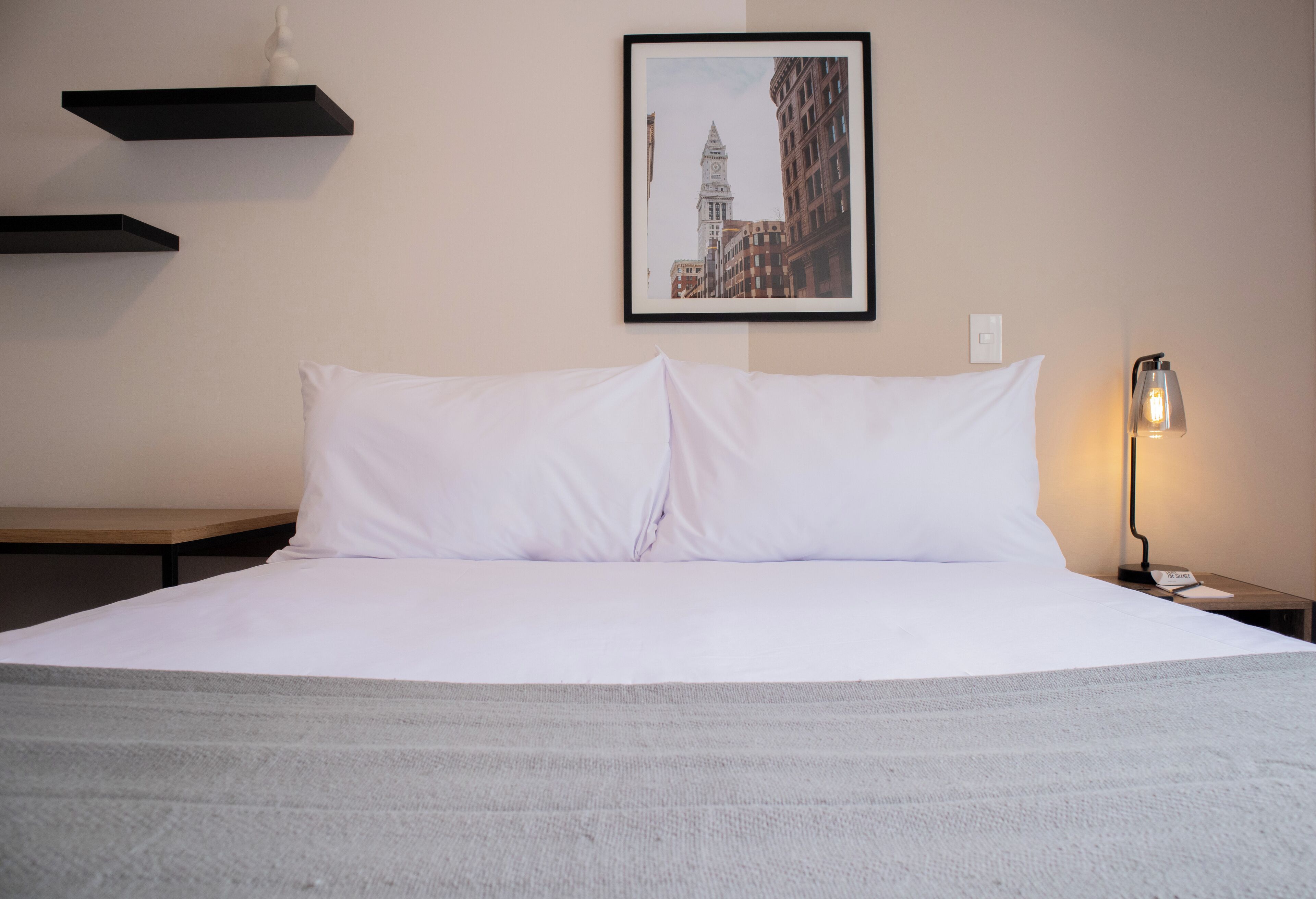 A hotel room with a large bed, a framed picture of a building on the wall, a bedside lamp, and a small table.