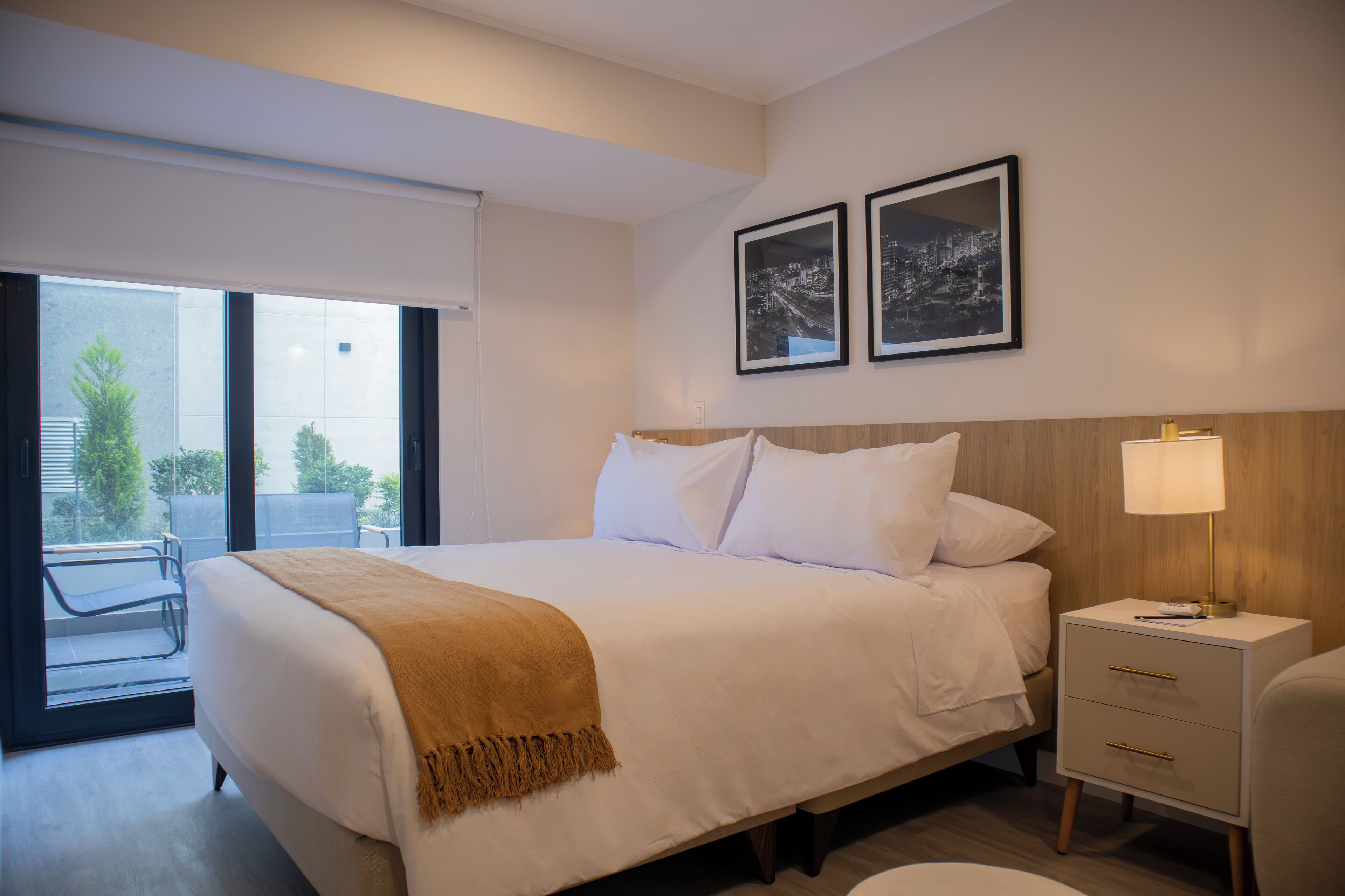A modern hotel room with a large bed, a bedside table with a lamp, and a view of the outdoors through a sliding door.