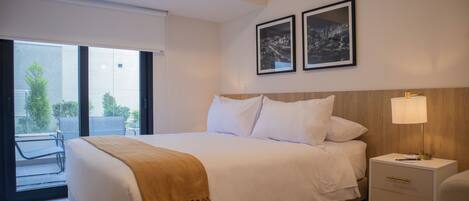 A modern hotel room with a large bed, a bedside table with a lamp, and a view of the outdoors through a sliding door.