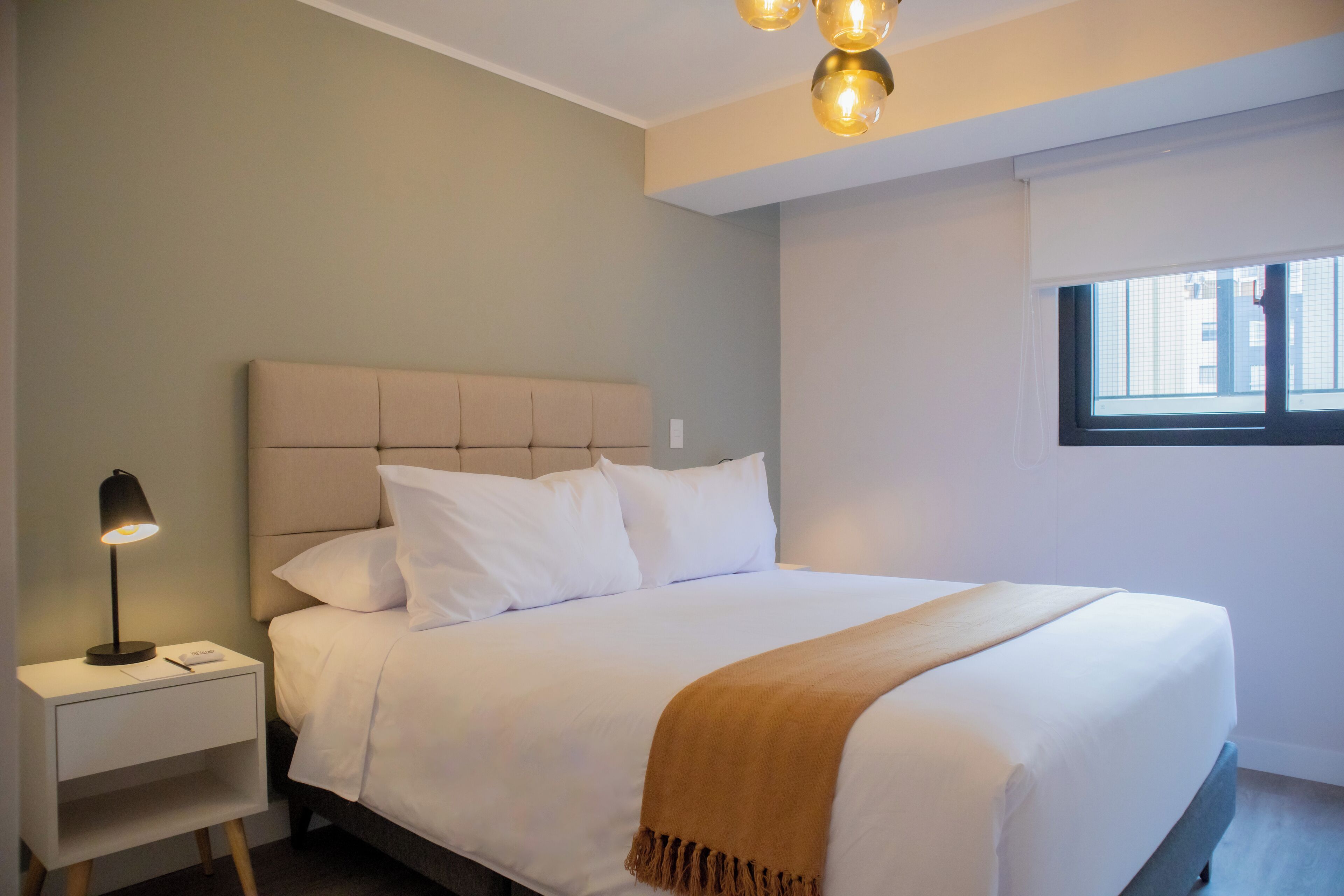 A neatly made bed with a headboard, a bedside lamp, and a window with blinds.