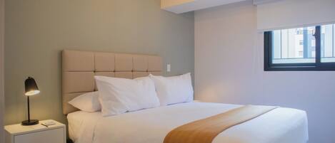 A neatly made bed with a headboard, a bedside lamp, and a window with blinds.