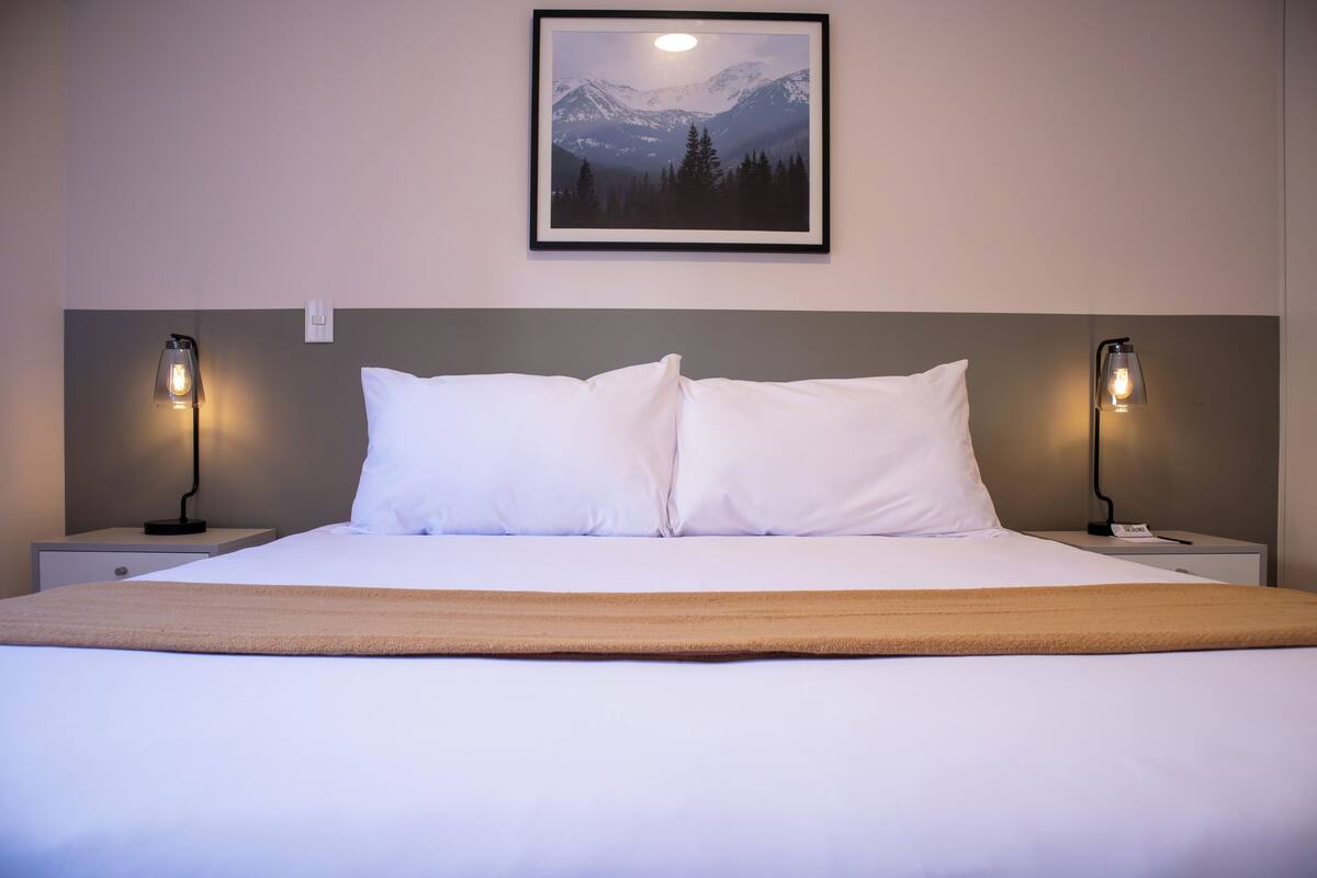 Ein ordentlich bezogenes Bett mit weißen Bettwäsche, zwei Nachttischlampen und ein gerahmtes Bild einer Berglandschaft an der Wand.