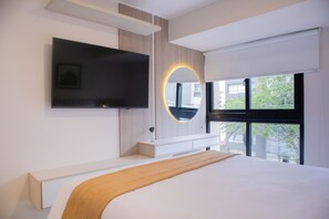 Une chambre moderne avec un grand lit, un téléviseur fixé au mur, une table de chevet et une fenêtre donnant sur des immeubles et des arbres.