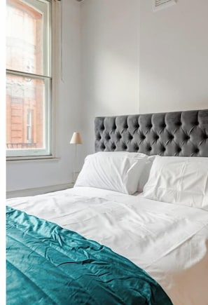 Uma cama bem arrumada com roupa de cama branca e azul-turquesa, uma cabeceira cinza com detalhe em botão e uma janela com vista para um edifício de tijolos.