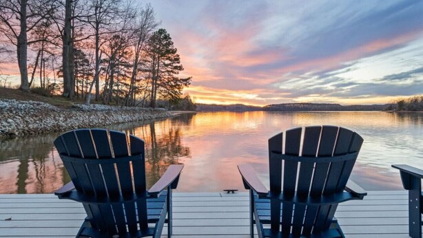 Dos sillas Adirondack mirando un lago tranquilo al atardecer, con árboles a la izquierda.