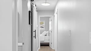 Un pasillo estrecho con paredes y puertas blancas que dan a un dormitorio con cama y ventana.