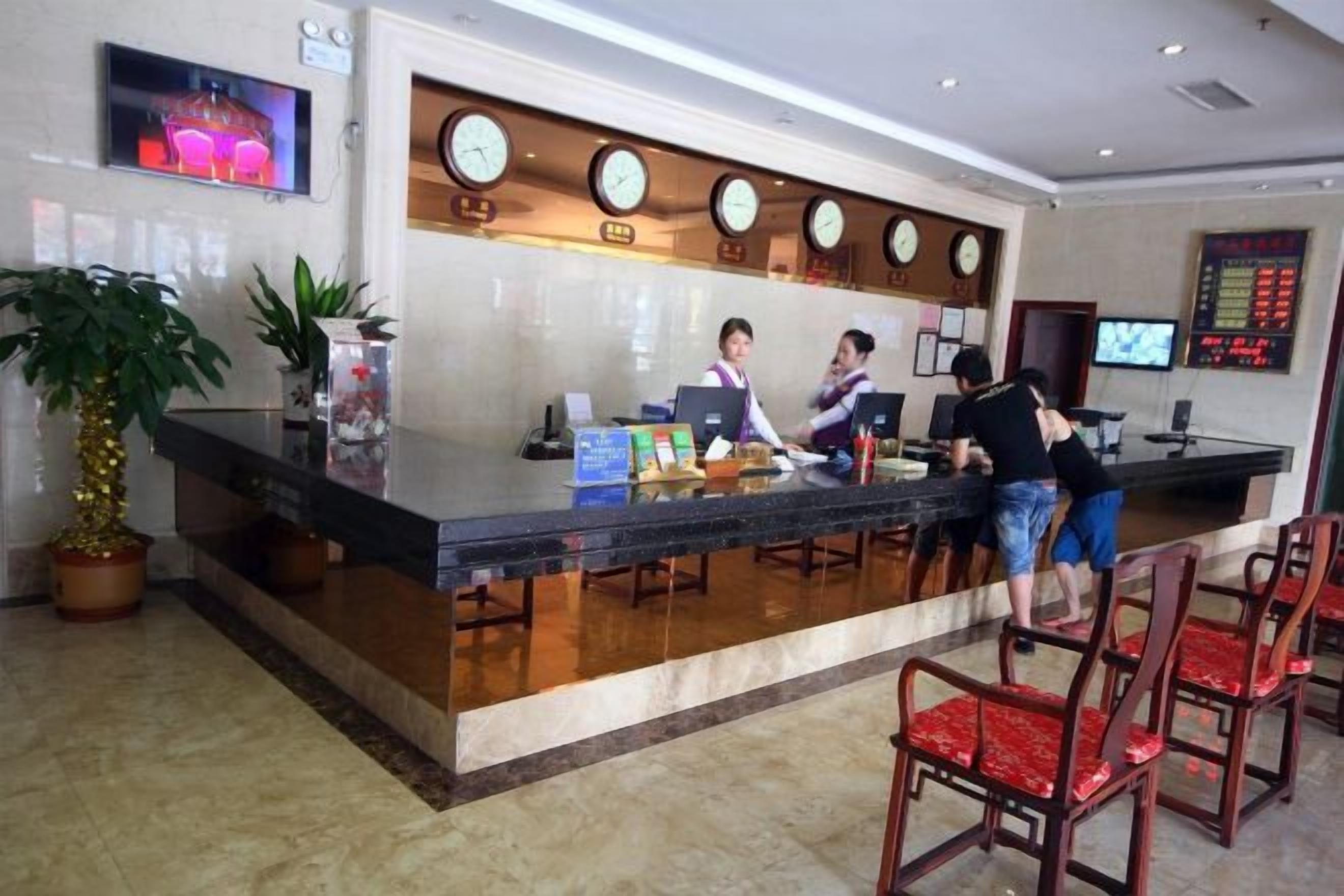 A hotel reception area with a counter, two receptionists, a TV screen, and a clock.