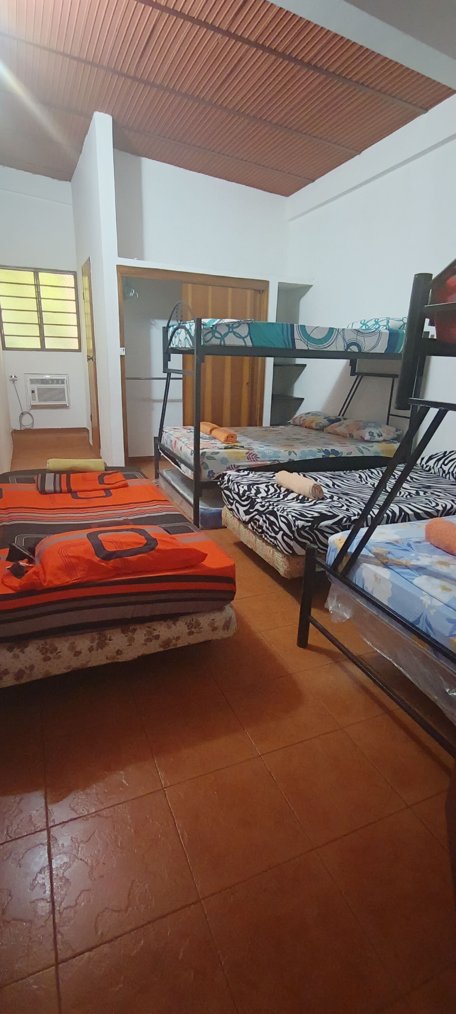 A dormitory room with bunk beds, a window with bars, a radiator, and a wooden ceiling.