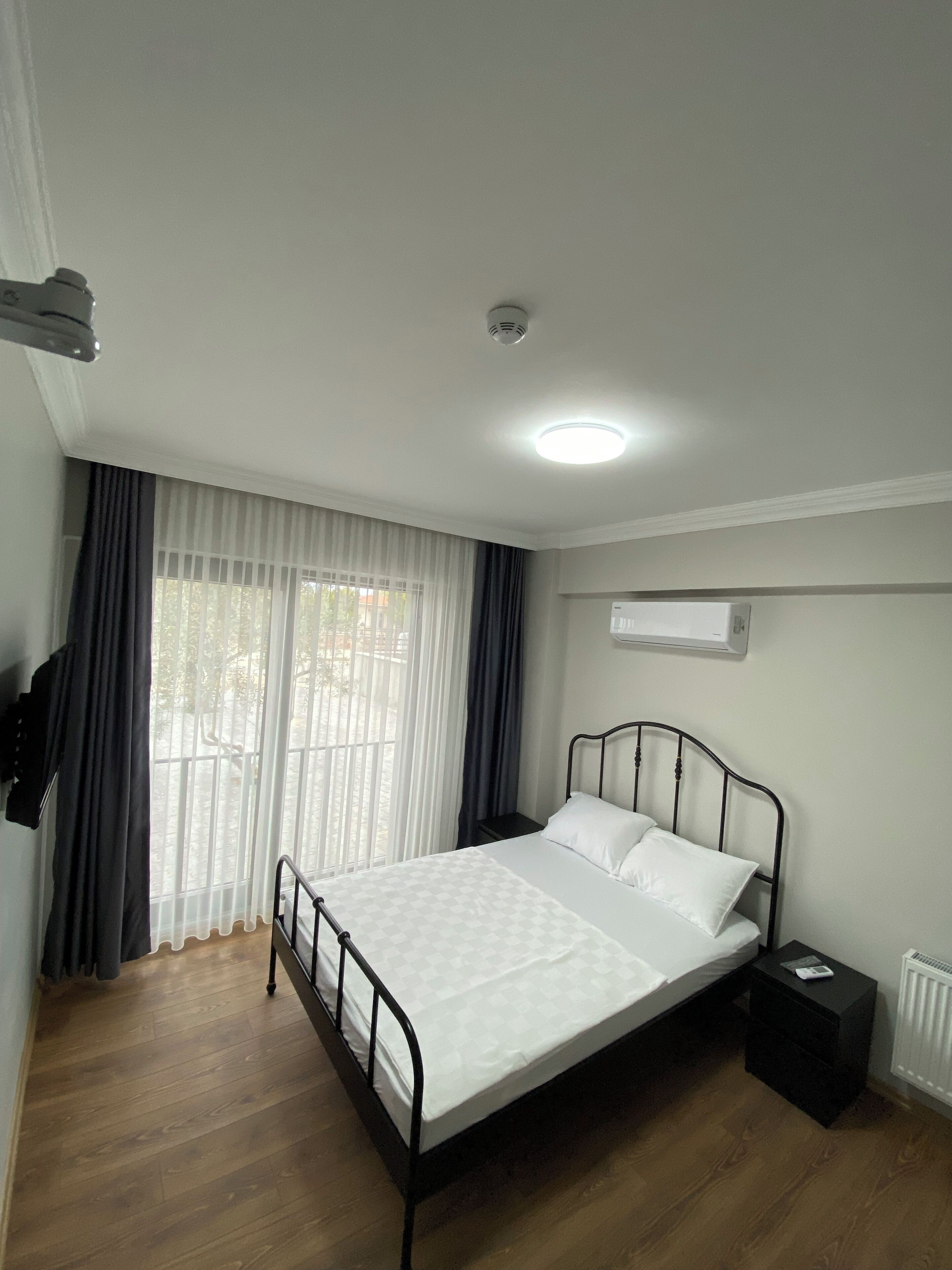 Un dormitorio con cama, una mesita de noche, ventana con cortinas, lámpara en el techo y un equipo de aire acondicionado.