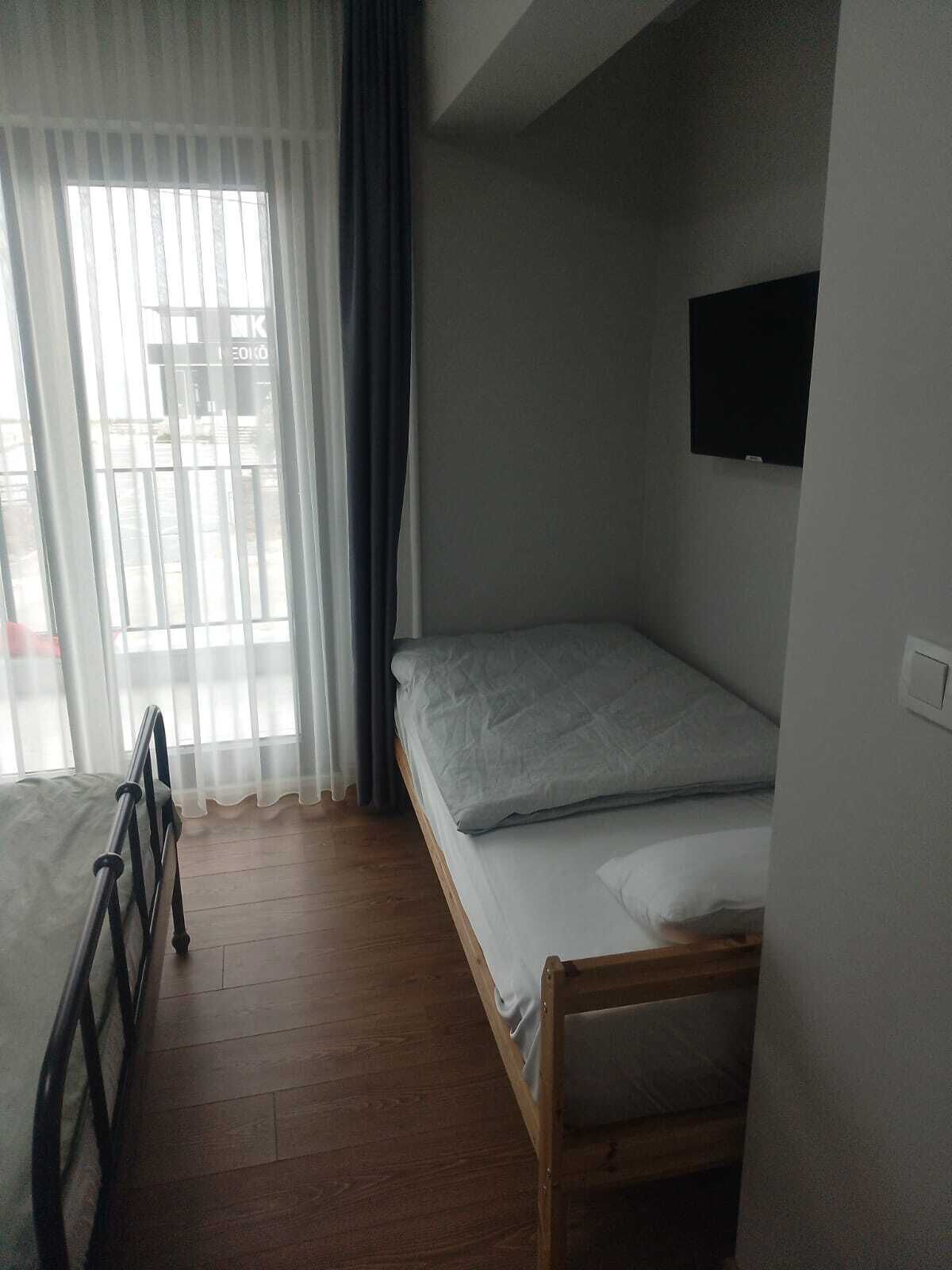 Una habitación pequeña con cama individual, suelo de madera, ventana con cortinas translúcidas y televisor montado.