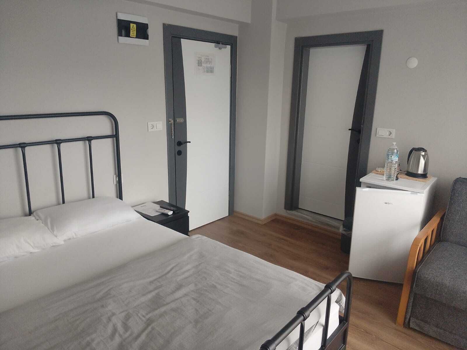 A hotel room with a bed, a nightstand, a chair, and a small table with a kettle and water bottle.