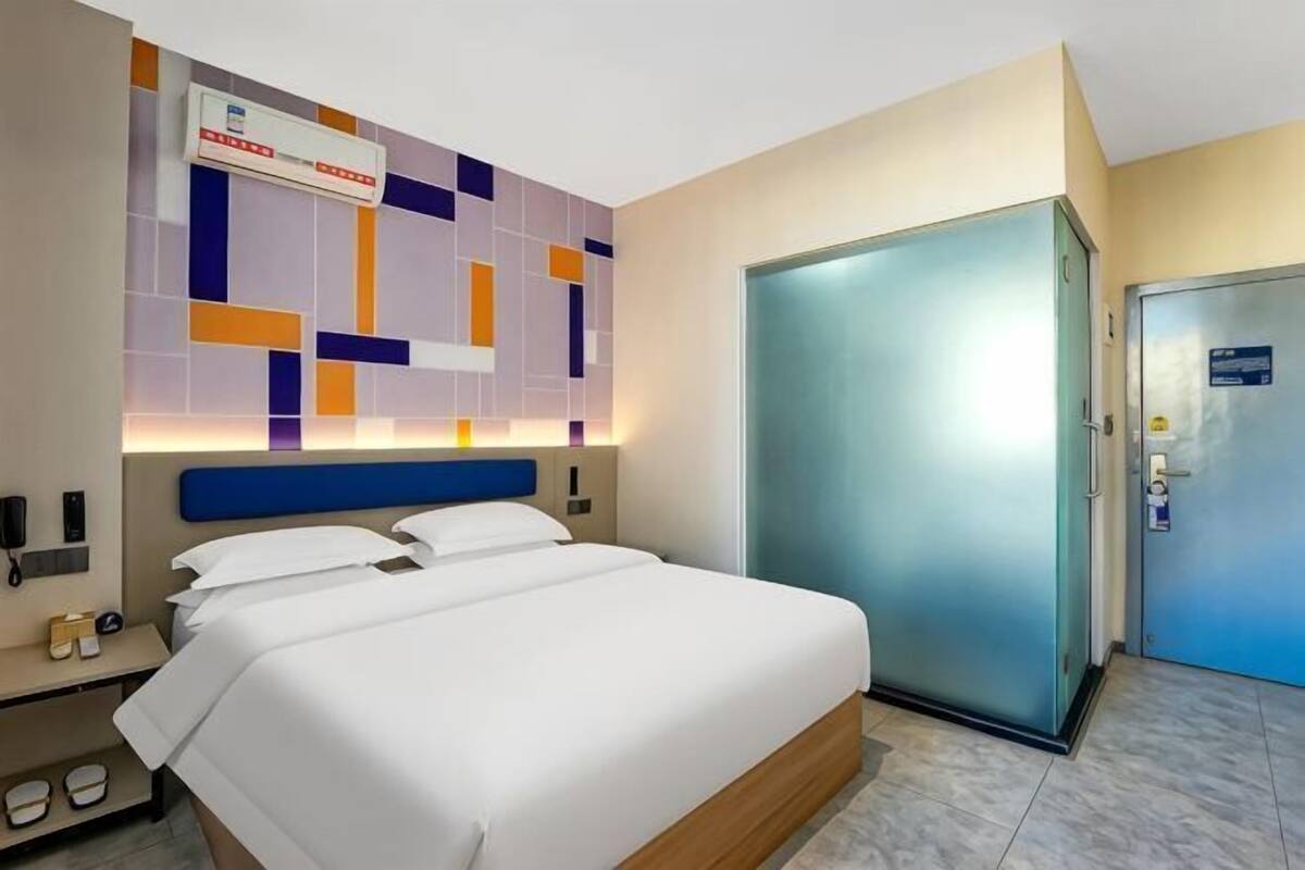 A hotel room with a bed, a headboard, a wardrobe, a nightstand, and a wall-mounted air conditioning unit.