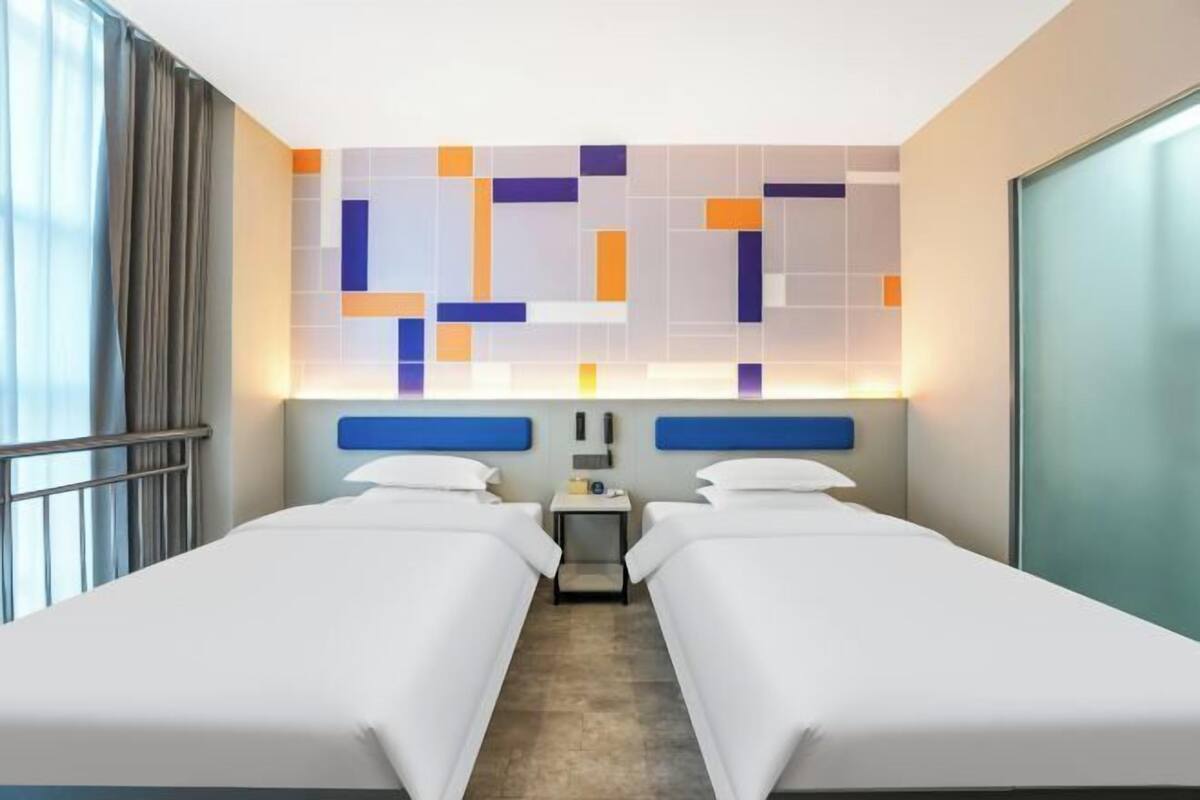 A hotel room with two beds, a small bedside table, a wall-mounted TV, and a window with curtains.