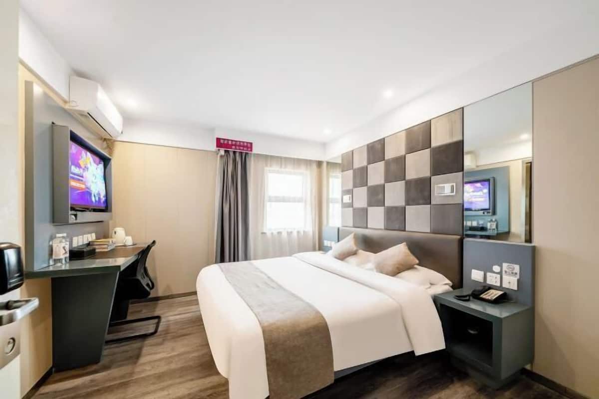 A modern hotel room with a large bed, a desk with a flat-screen TV, a small bedside table, and a large mirror.