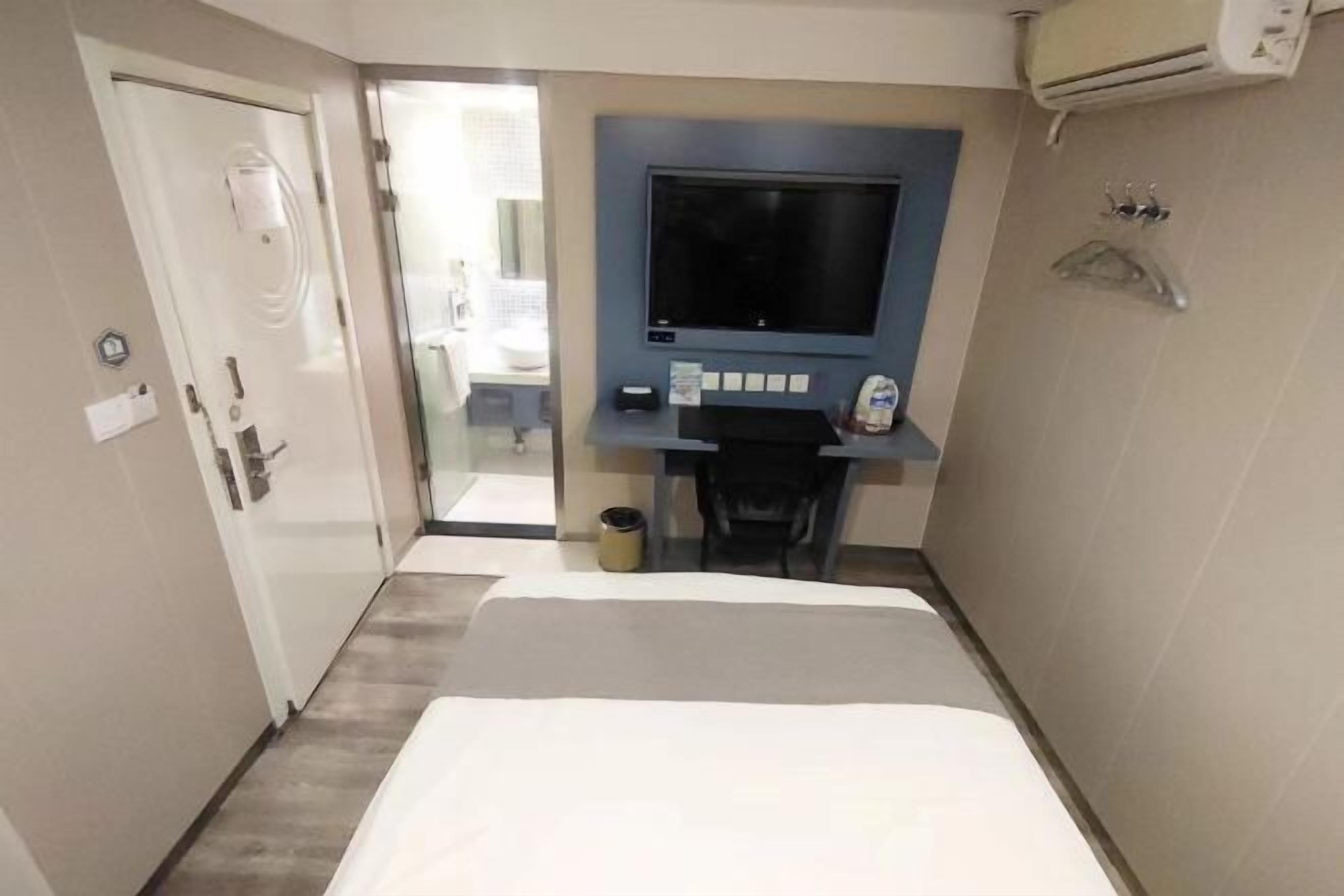 A hotel room with a bed, a desk with a flat-screen TV, a chair, and a small trash bin.