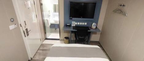 A hotel room with a bed, a desk with a flat-screen TV, a chair, and a small trash bin.