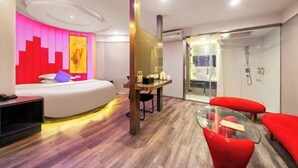 Una habitación de hotel moderna con una cama curva, cabina de ducha de vidrio y una pequeña zona de estar con sillas rojas.