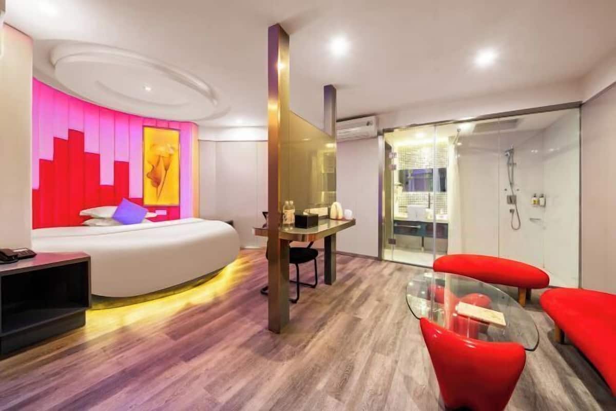 A modern hotel room with a curved bed, a glass shower enclosure, and a small seating area with red chairs.
