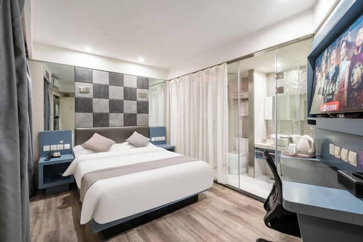 A modern hotel room with a large bed, a TV, a desk, and a bathroom visible through a glass partition.