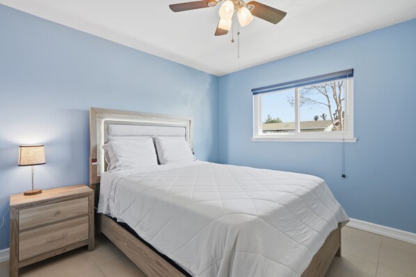 A bedroom with a bed, nightstand, lamp, ceiling fan, and window with blinds.