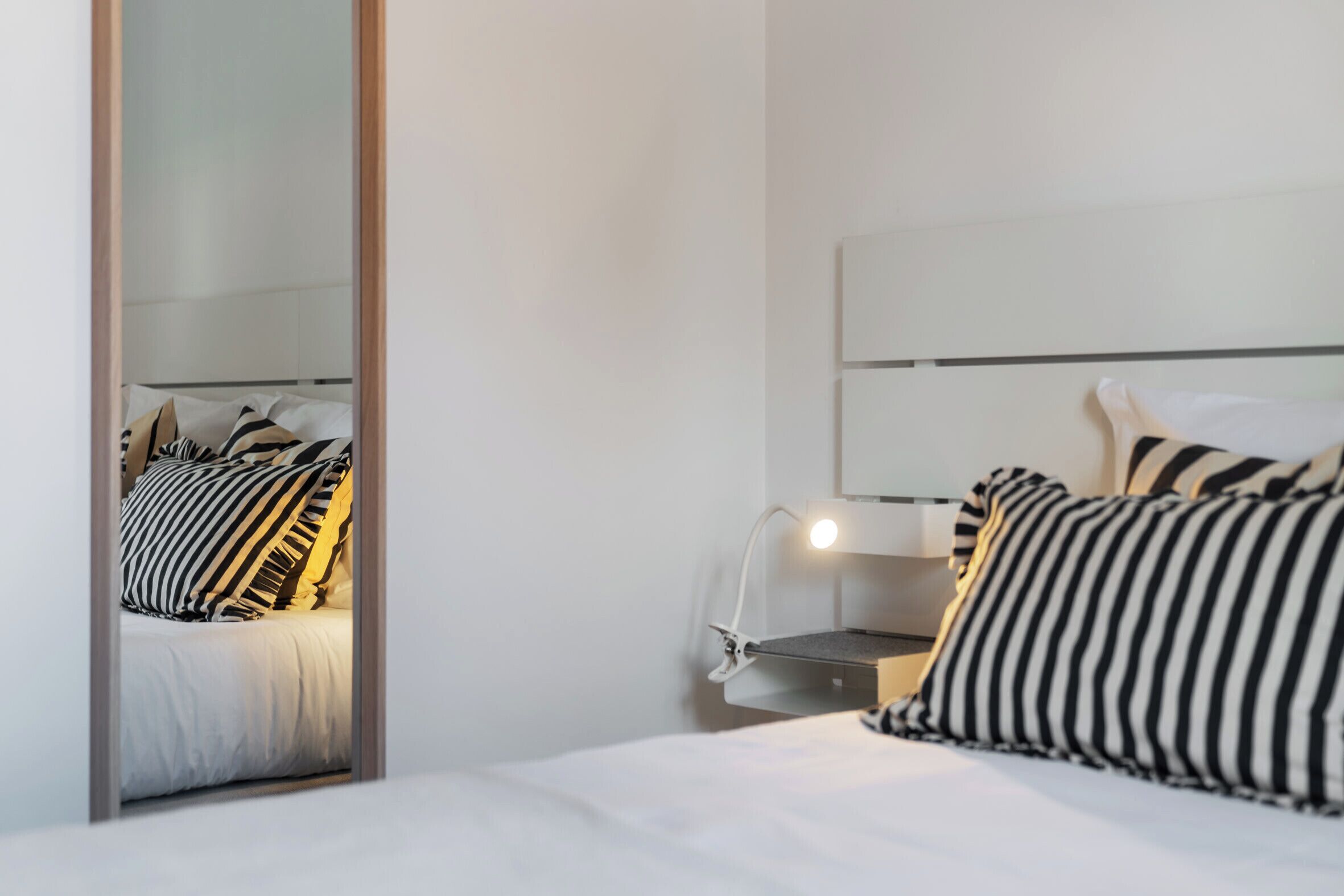 A modern hotel room with a bed, a bedside table with a lamp, and a mirror reflecting the bed.