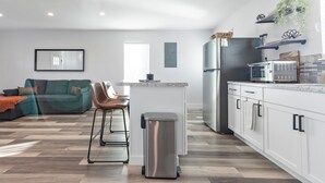 Eine moderne Küche mit weißen Schränken, einem Edelstahl-Kühlschrank und einer Mikrowelle. Im Vordergrund steht ein Barhocker und ein Mülleimer.